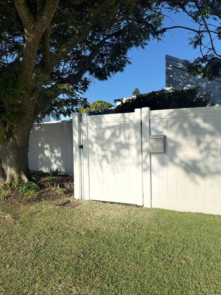 White composite fence with gate running along a grassy yard with tree overhead and residential building in background