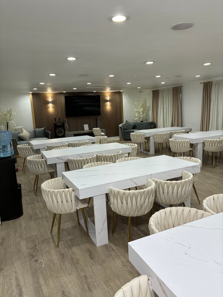 Long, bright view of our event space with white marble tables, tufted cream chairs, recessed lighting, and a wooden accent wall with bronze fixtures
