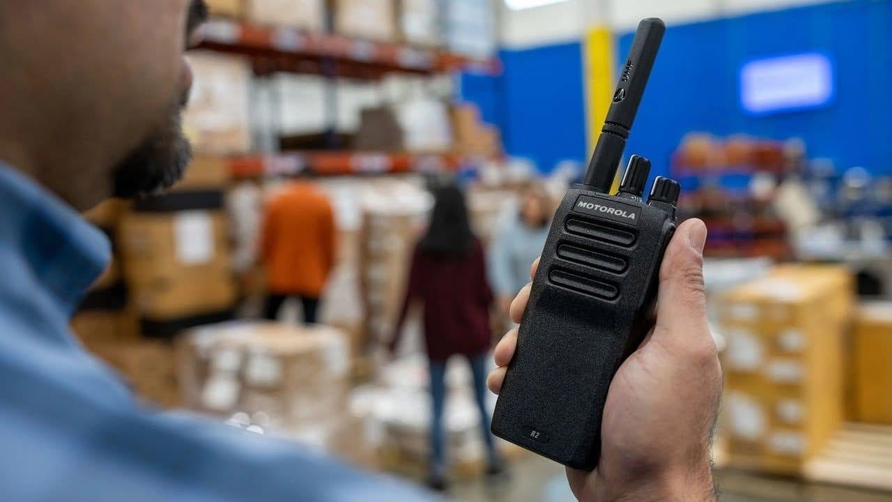 Worker holding a black two-way radio in a warehouse with blue wall and shelving in background