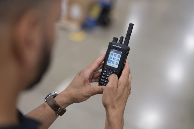 Person holding a black handheld two-way radio with antenna, displaying the screen and keypad, in an indoor setting