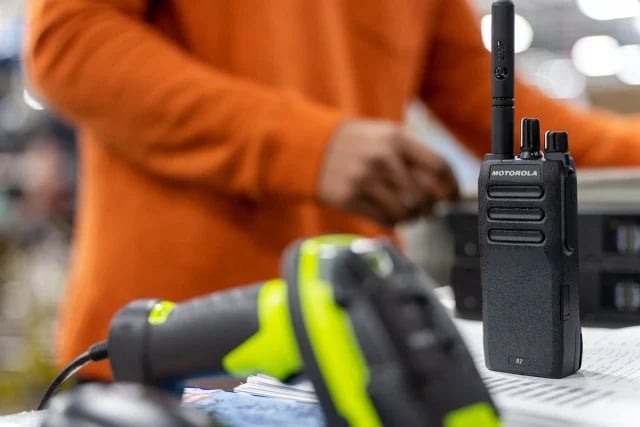 Worker in orange jacket holding black Motorola two-way radio with yellow safety gloves in foreground on outdoor work table