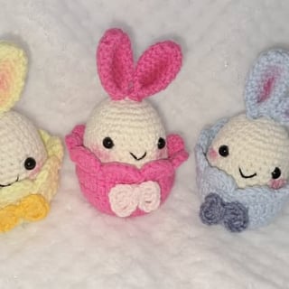 Three crocheted amigurumi bunnies in pink, yellow, and blue with bows, displayed on white fabric