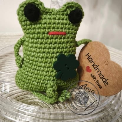 Handmade crocheted green frog amigurumi with black eyes and red mouth, displayed with a kraft paper heart tag
