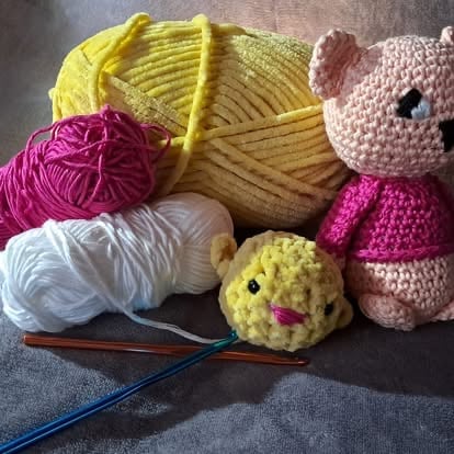 Colorful crochet supplies including yarn balls in pink, yellow, and white, two handmade amigurumi animals, and a crochet hook on gray background