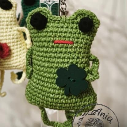 Green crocheted frog keychain with black eyes, pink mouth, and a dark green clover charm on its body
