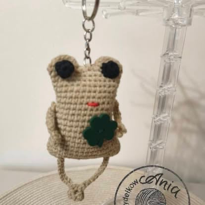 Small crocheted beige frog keychain with black eyes, red nose, and green heart detail hanging from a chain on a decorative glass stand