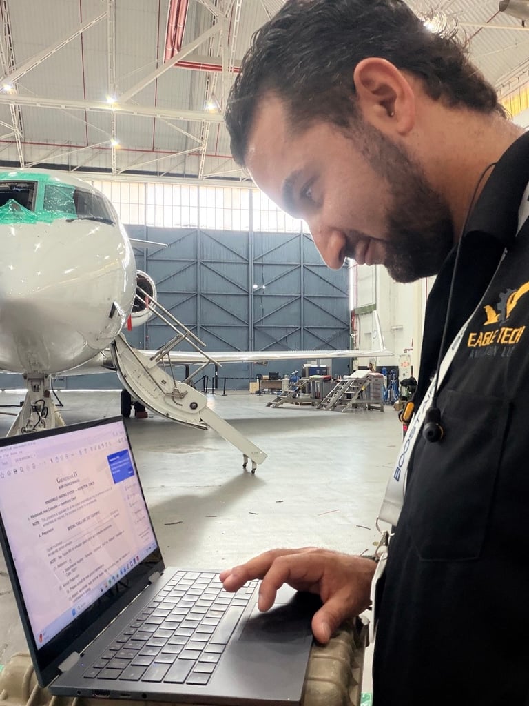 Bearded technician working on laptop inside aircraft hangar with large passenger airplane in background