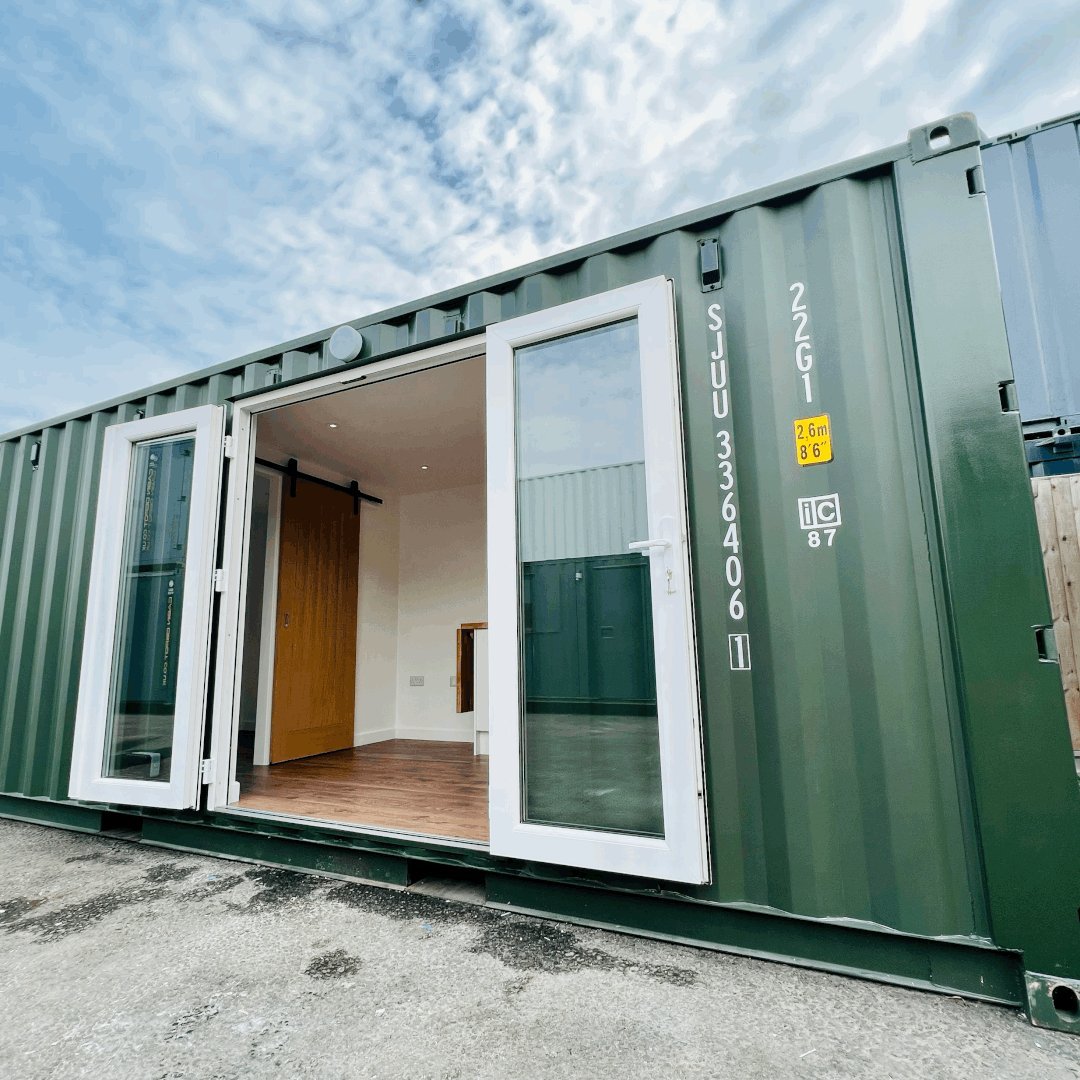 Modern green shipping container home with open white doors revealing wood flooring and minimalist interior design