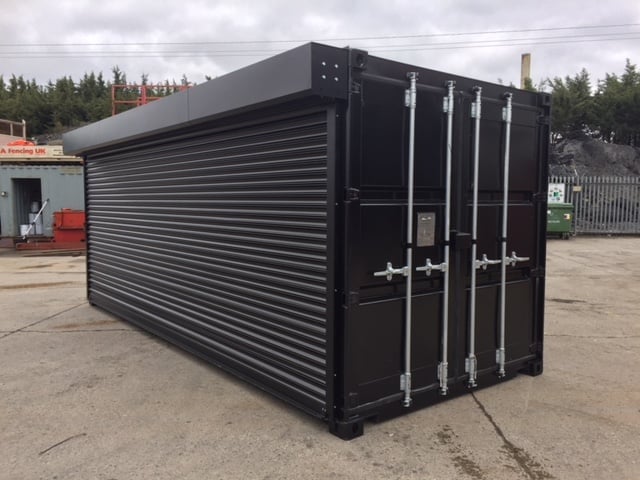 Black corrugated metal shipping container with double doors in a commercial outdoor lot surrounded by trees and buildings