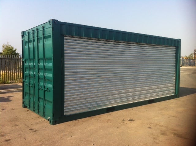 Green shipping container with metal roller shutter door on sandy ground in an open lot