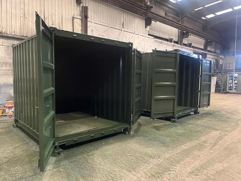 Three open green shipping containers lined up in an industrial warehouse with corrugated metal ceiling