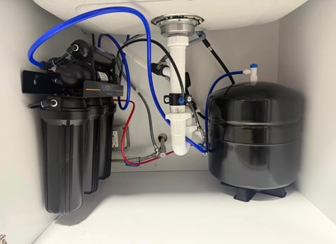 Under-sink water filtration system with black filter housing, blue tubing, and metal storage tank mounted in cabinet.