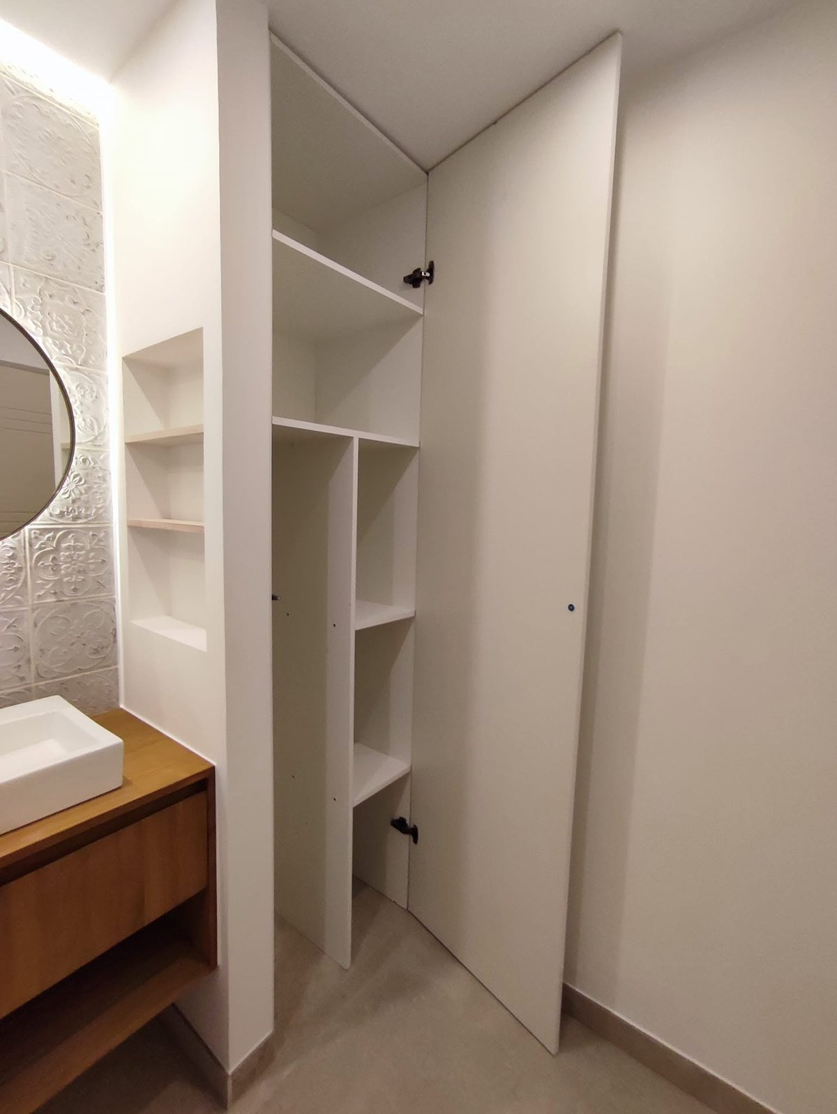 Moderner weißer Einbauschrank mit geöffneter Tür und verstellbaren Einlegeböden, angrenzend an ein Badezimmer mit Holzwaschtisch und rundem Spiegel
