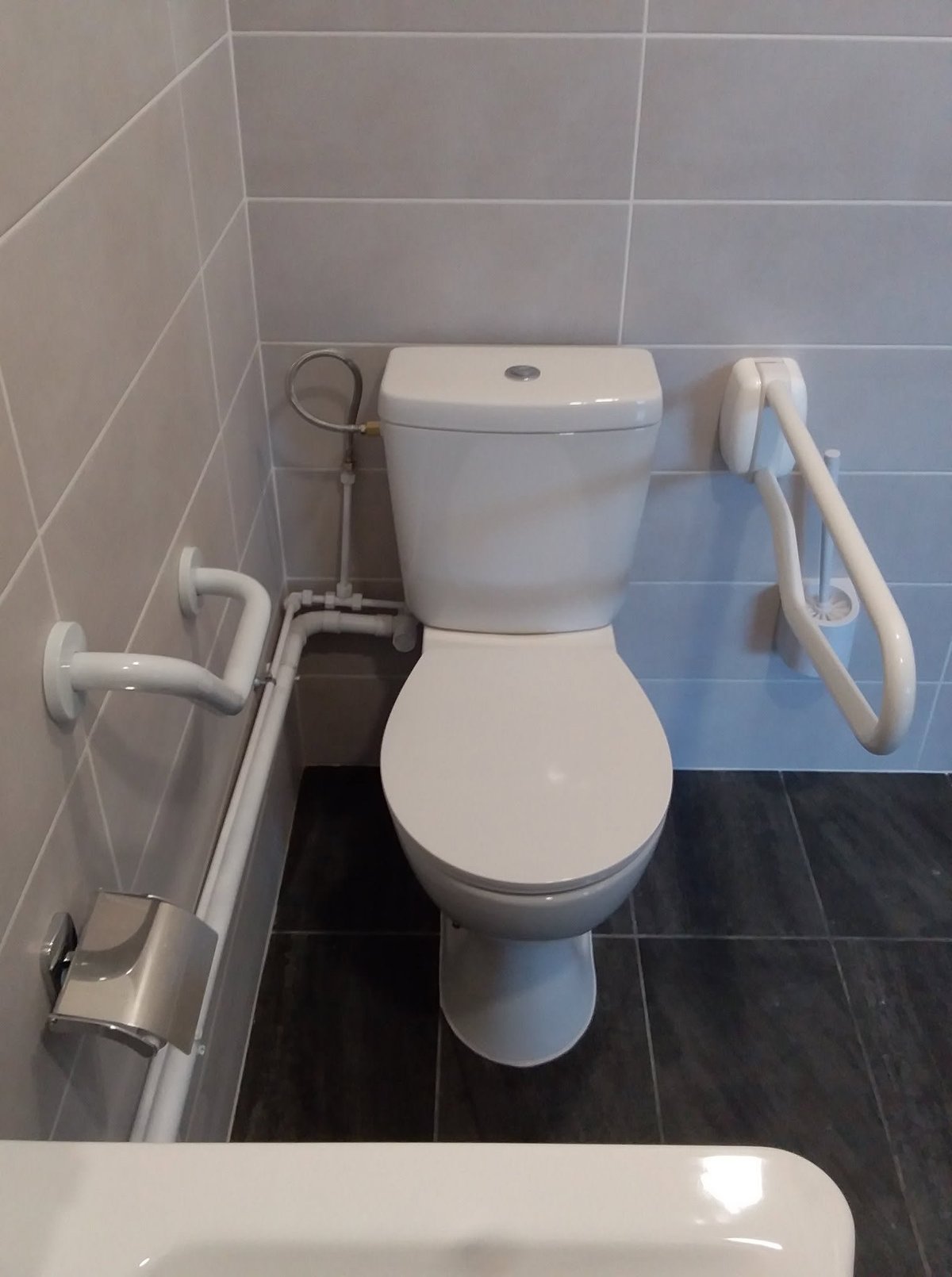 Compact bathroom with white two-piece toilet, gray tile walls, and black floor tiles. Toilet paper holder and bathroom accessories visible.