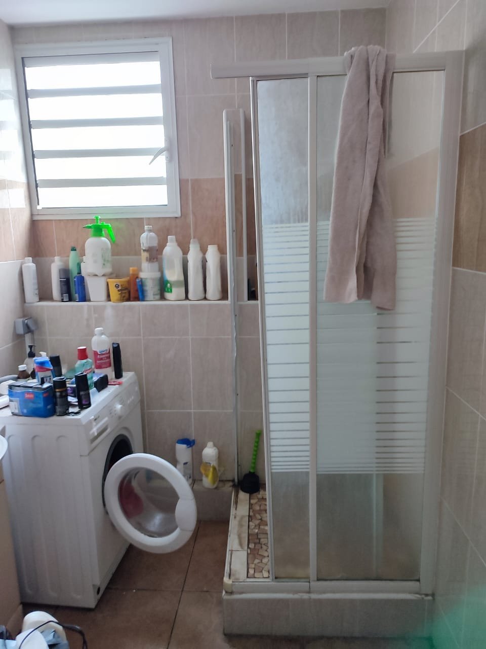 Compact bathroom with washing machine, glass shower enclosure, beige tiles, and shelving with toiletries and supplies