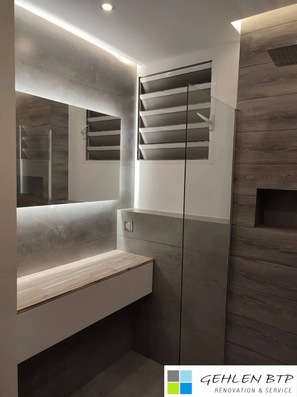 Modernes Badezimmer mit beleuchtetem Spiegel, weißem Waschtisch und glasabgetrennter Dusche mit holzartigen Fliesenakzenten