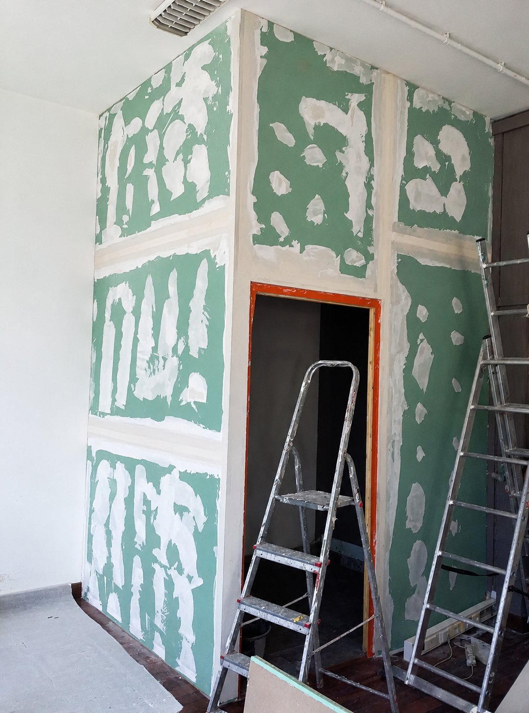 Trockenbauwand im Bauzustand mit grünen Gipskartonplatten, verspachtelten Fugen, orangem Durchgangsrahmen und Aluleitern