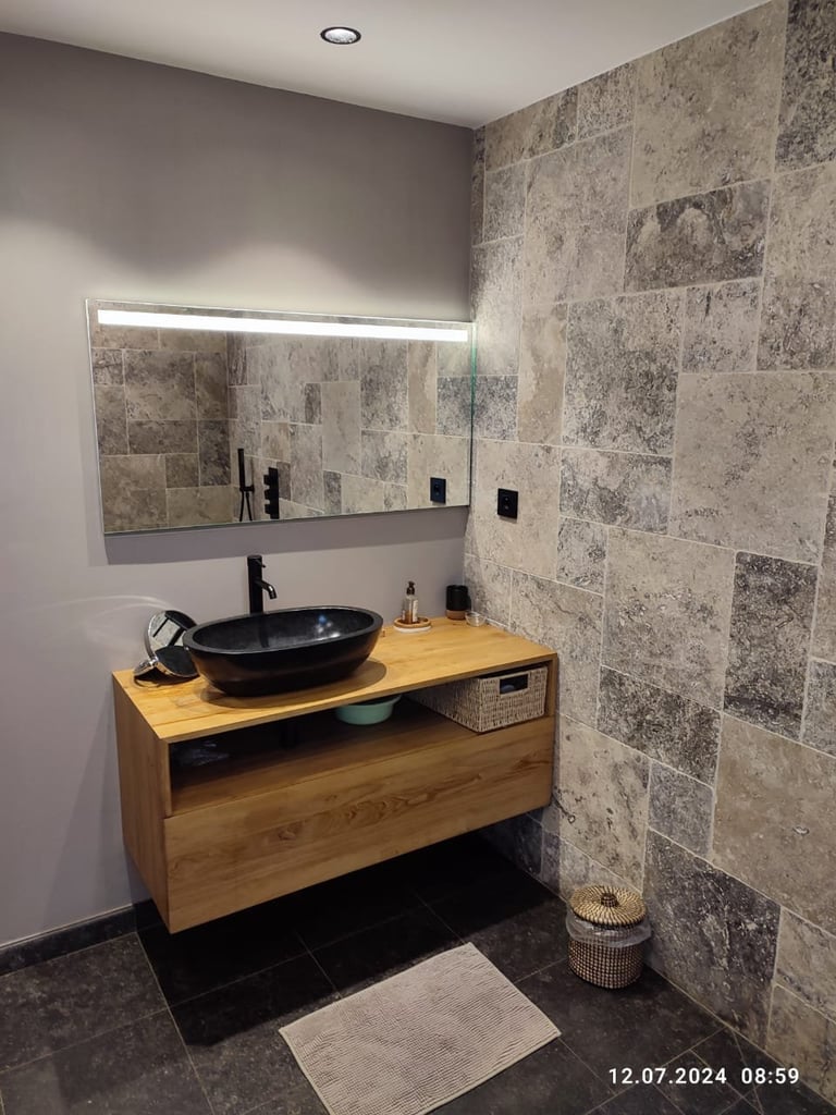 Modern bathroom with wooden vanity, black round sink, tile walls, illuminated mirror, and black fixtures