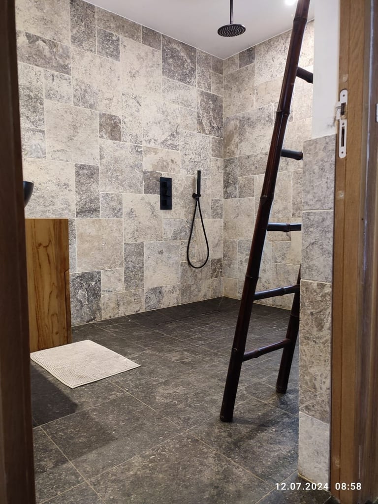 Modern walk-in shower with beige and gray tiled walls, dark floor, overhead rainfall showerhead, handheld sprayer, and wooden ladder towel rack
