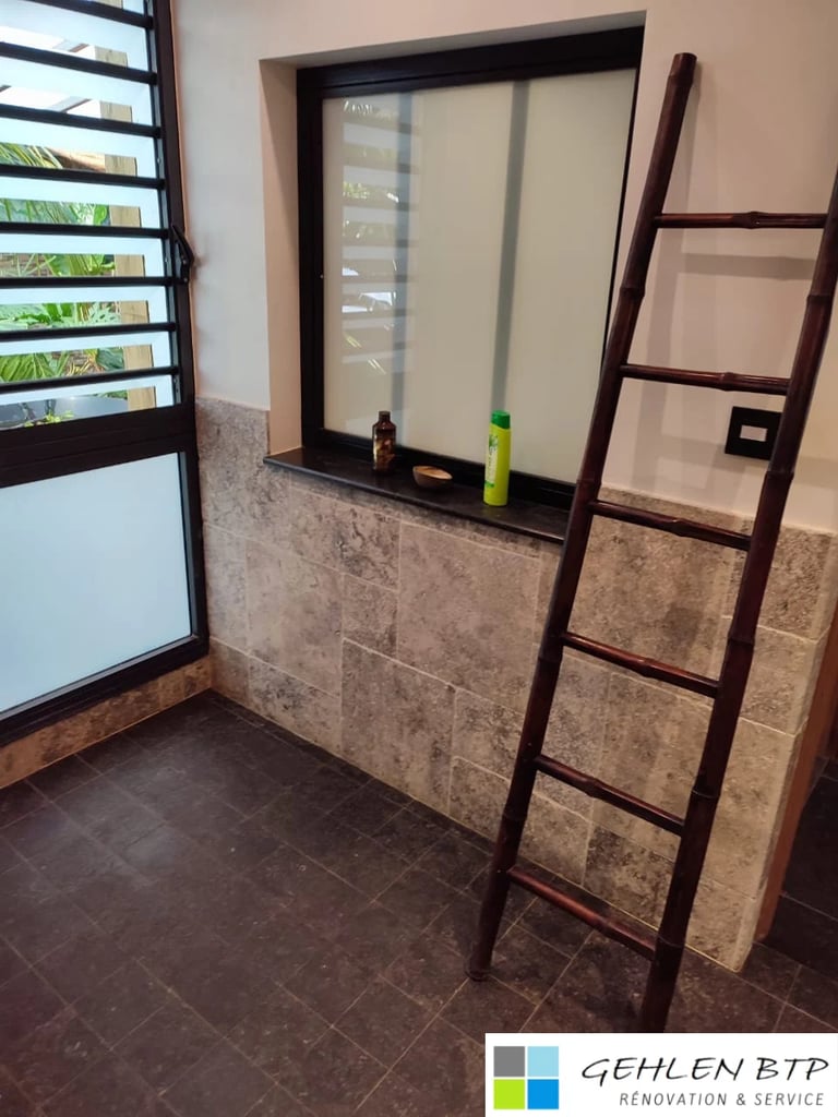 Interior bathroom space with black-framed mirror, wooden ladder, tile walls, and louvered window with plant visible outside