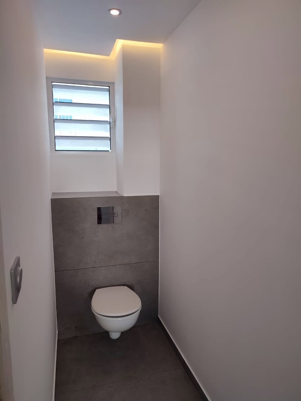 Modernes Badezimmer mit wandhängendem weißem WC, dunkler Fliesenwand, horizontalem Fenster mit Jalousie und warmer Deckenbeleuchtung