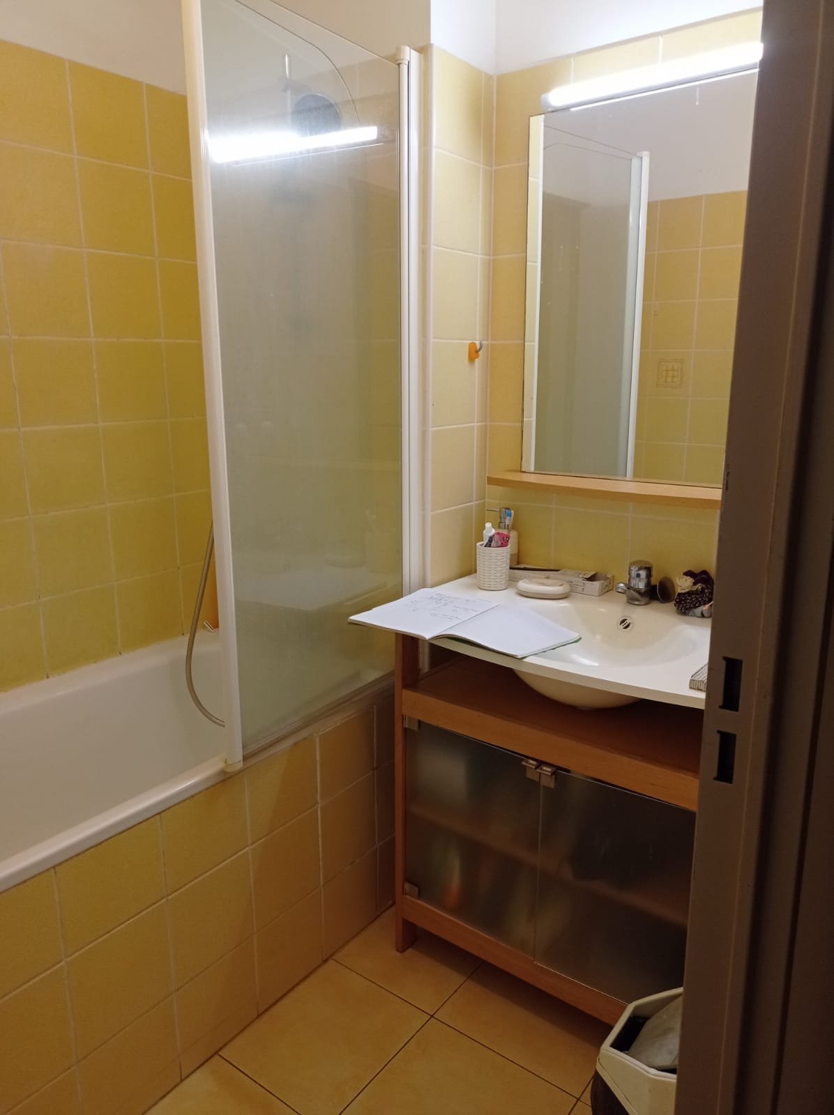 Vorher — Badezimmer mit gelben Fliesenwänden, Glasduschabtrennung, weißem Waschbecken, Spiegel mit Beleuchtung und Holzwaschtisch