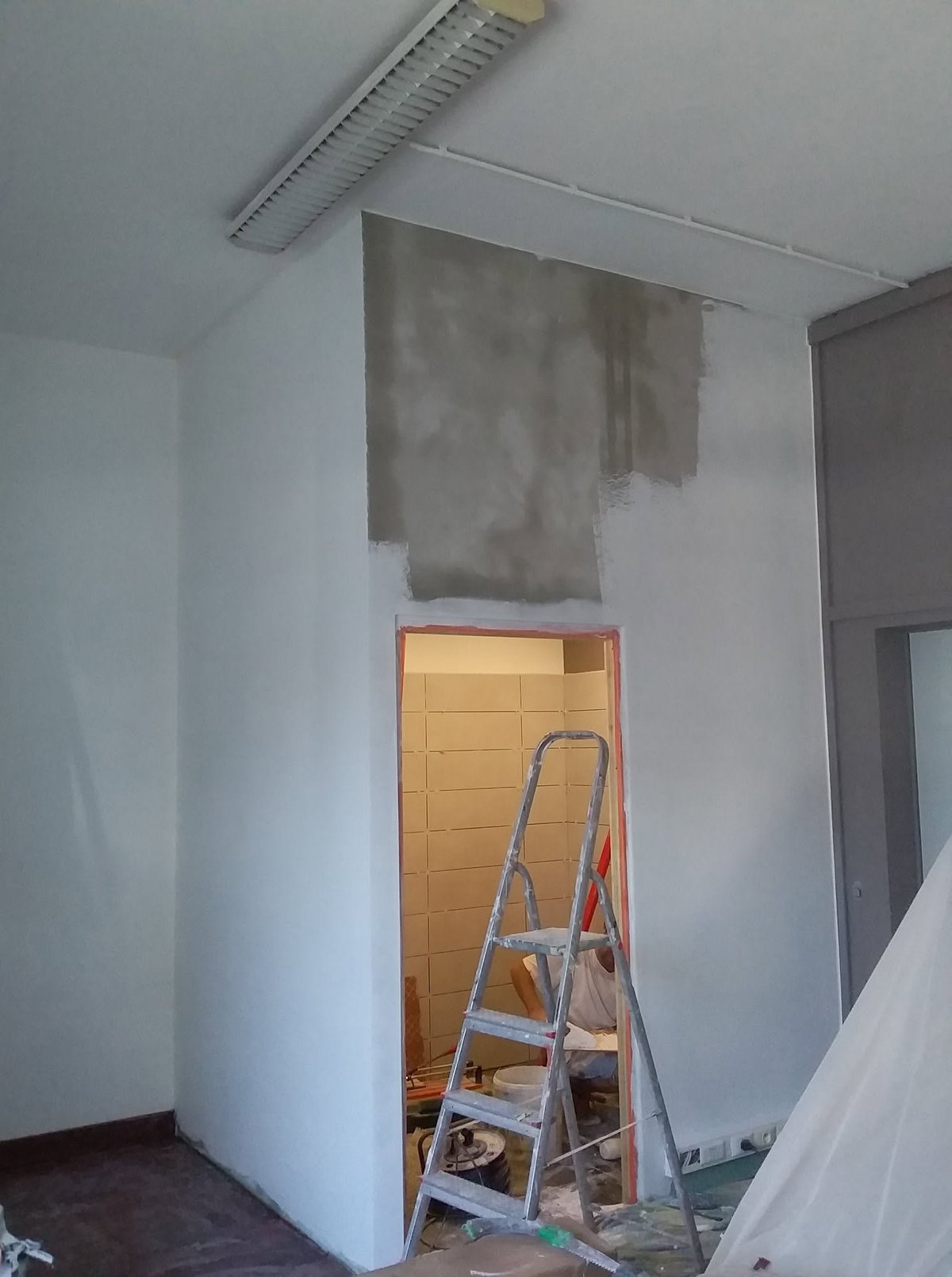 Interior room under renovation with white walls, metal ladder, plastic sheeting, and visible ceiling vents and construction materials