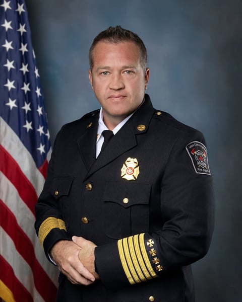 Portrait of a male fire chief in formal black uniform with gold insignia and badges, standing in front of an American flag against a blue background