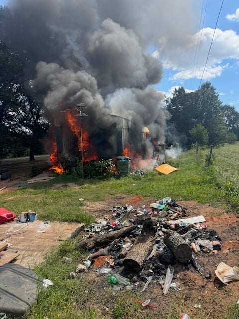 A structure engulfed in flames and heavy smoke with debris and burnt materials in the foreground, surrounded by overgrown grass and trees.
