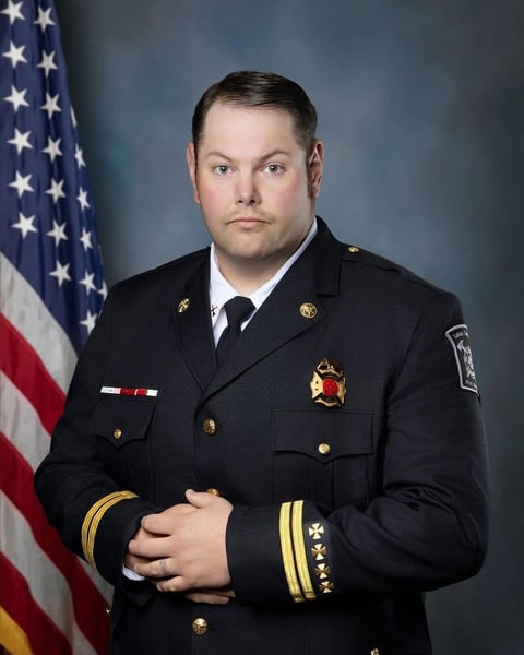 Firefighter in dress uniform with American flag in background, standing for official portrait