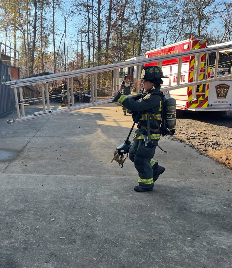 Firefighter in full gear walking toward fire truck at an outdoor training site with bare trees in background
