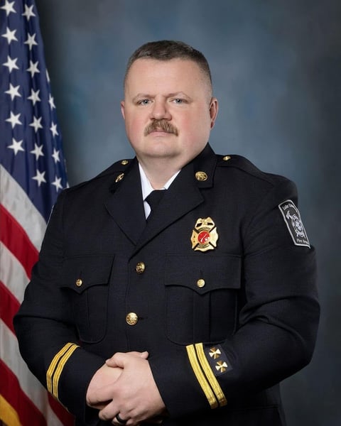 Male fire department official in dress uniform with insignia and medals, standing in front of American flag