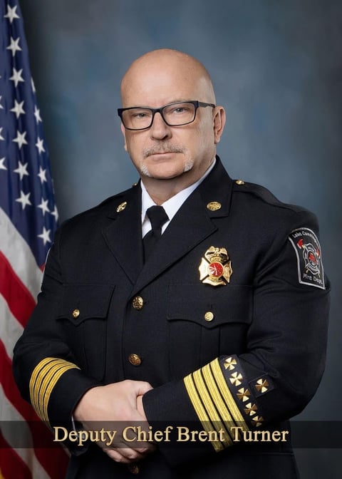 Official portrait of Deputy Chief Brent Turner in dark fire department uniform with gold insignia, standing before American flag.