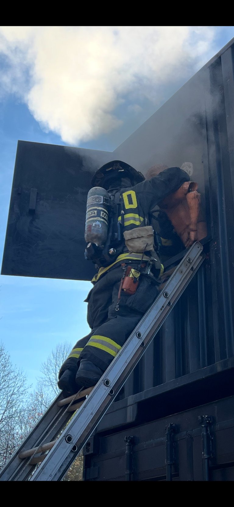 Firefighters in full gear climbing aluminum ladder against dark building with blue sky and clouds in background