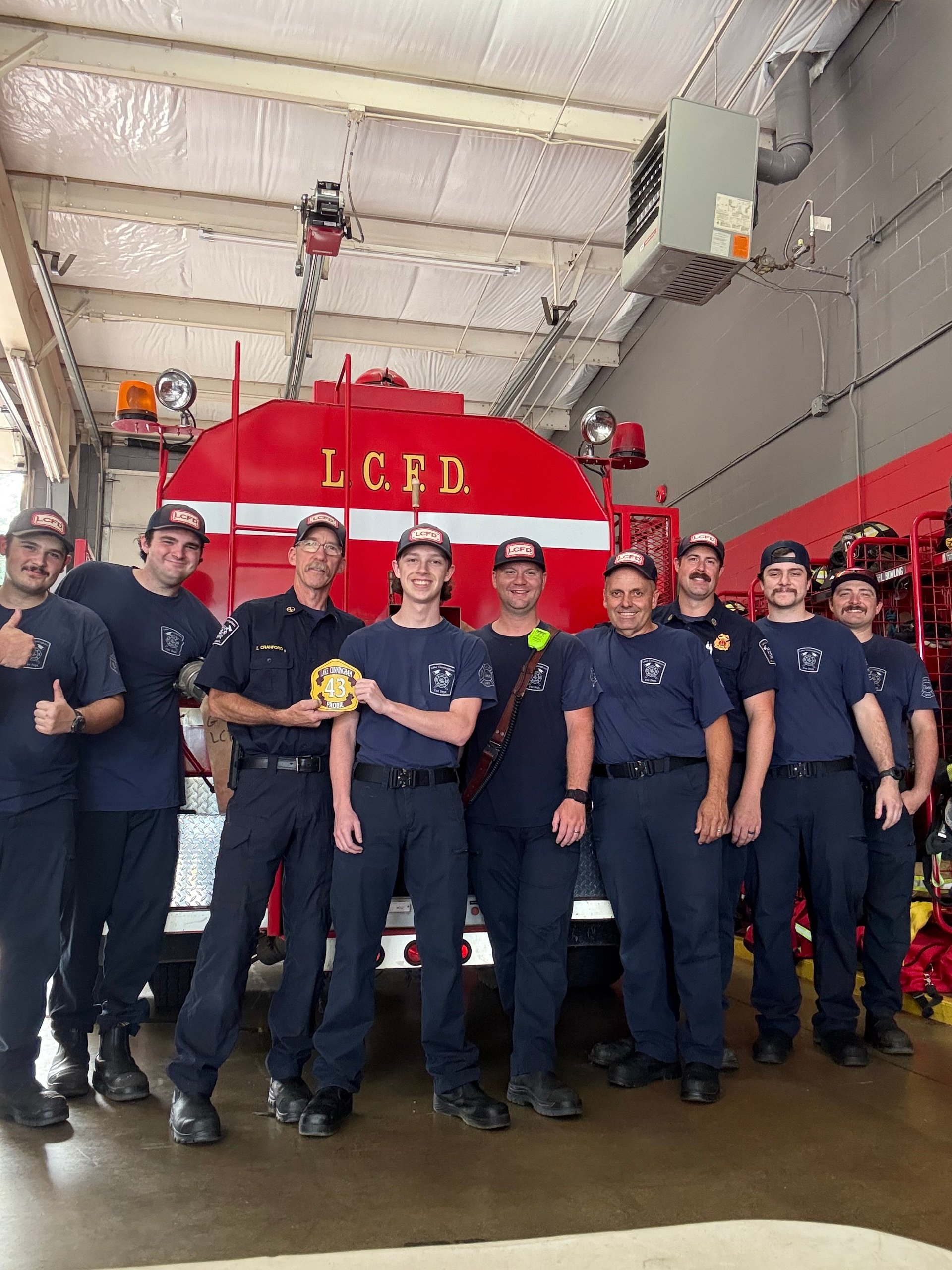 Group of firefighters in dark uniforms posing in front of a red fire truck marked LCFD in a station garage.