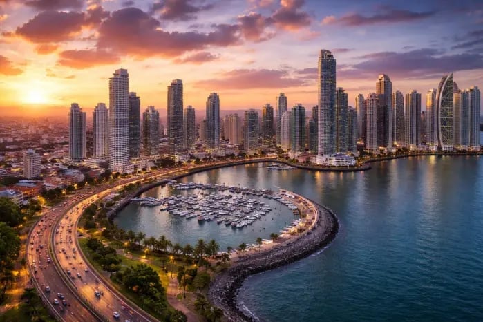 Aerial sunset view of a coastal city skyline with high-rise buildings, a curved marina filled with boats, and illuminated waterfront promenade