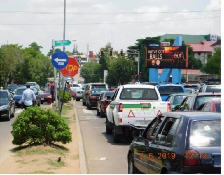 Busy street intersection with multiple parked cars, directional signs, storefronts with colorful signage, and green trees lining the roadside