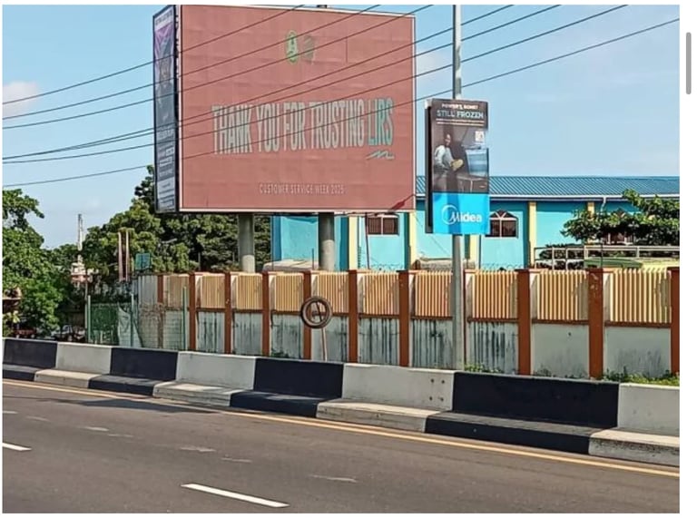Street view of a red billboard above corrugated metal fencing with blue storefront underneath and striped barrier posts along the road