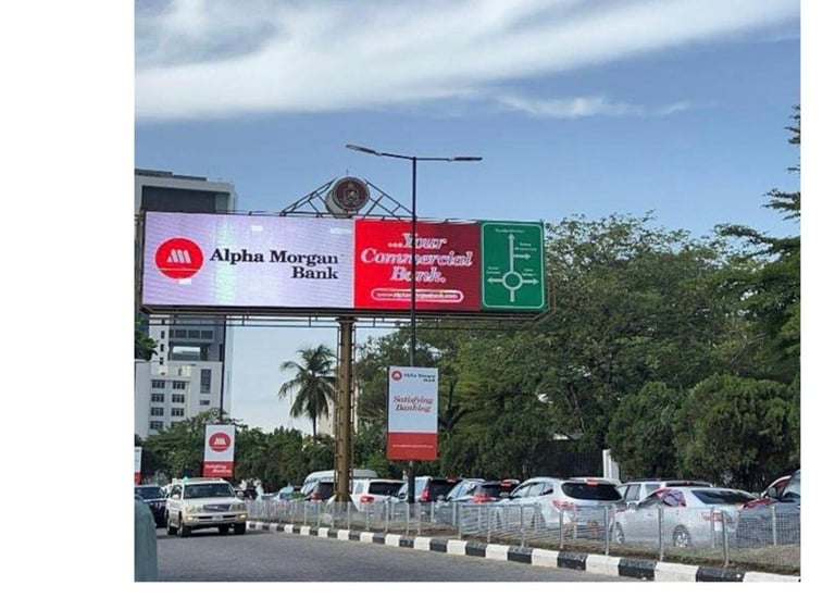 Large roadside digital billboards displaying Alpha Morgan Bank, Your Commercial Bank, and a medical clinic sign along Adetokunbo Ademola Street by Eko Hotel, with parked cars and a smaller Alpha Morgan Bank pole sign below
