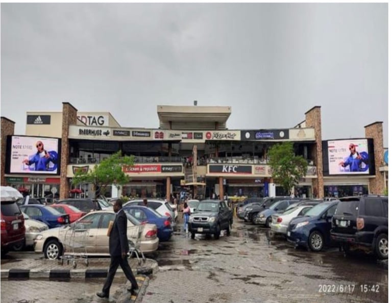 Shopping center parking lot with parked cars, digital billboards, and storefronts including KFC and other retail shops under cloudy sky