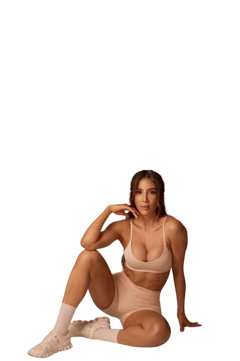 Woman in beige sports bra and high-waisted shorts sitting and posing with hand to head against white background