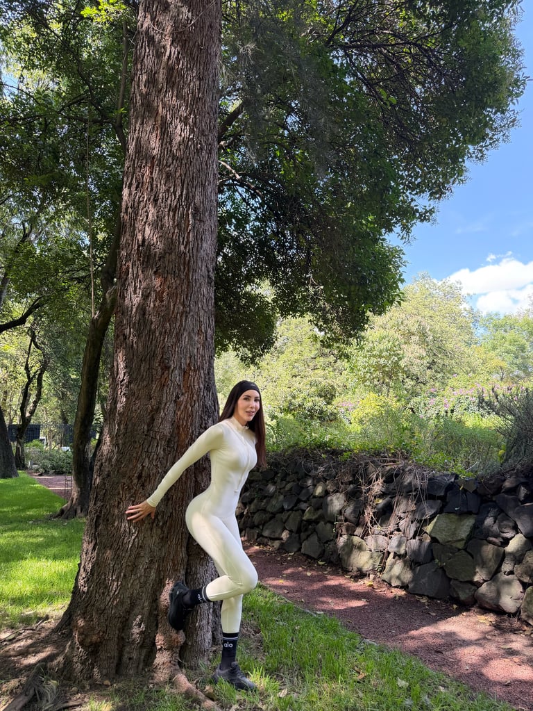 Woman in white athletic wear leaning against a large tree trunk in a park setting surrounded by greenery and a stone wall