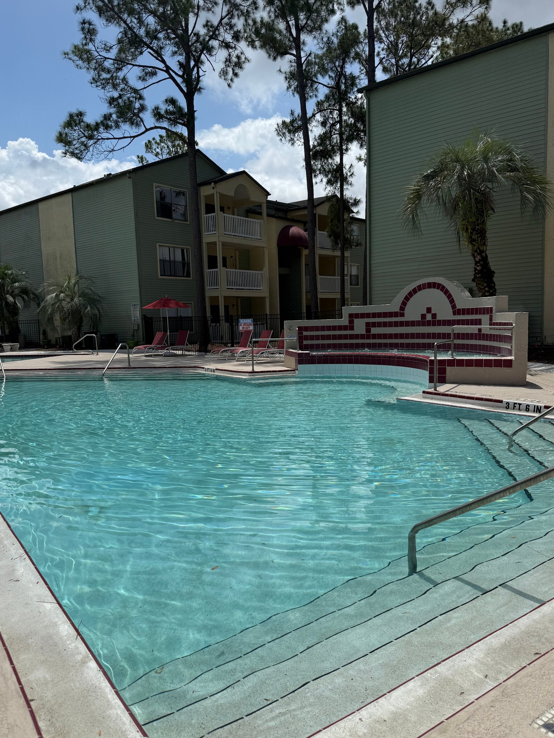 Apartment complex with turquoise swimming pool, lounge chairs, spa, and multi-story buildings surrounded by palm trees