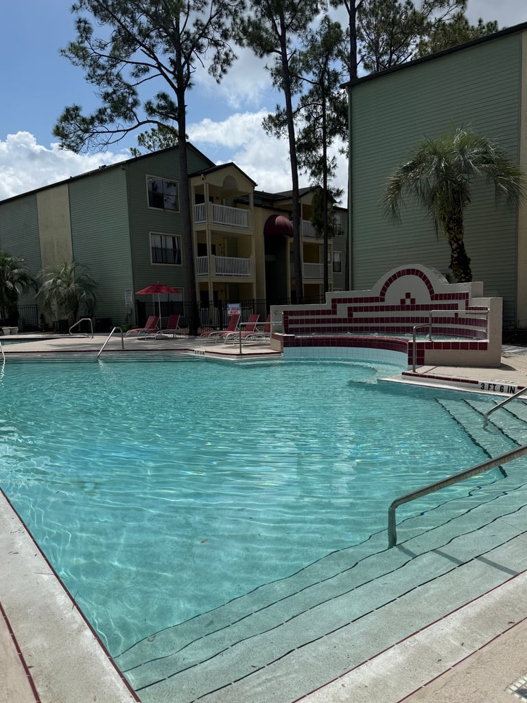Apartment complex with turquoise swimming pool, lounge chairs, spa, and multi-story buildings surrounded by palm trees