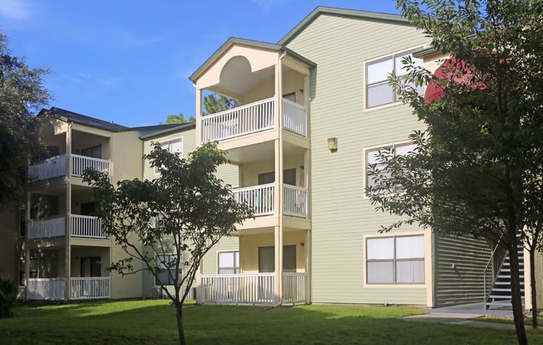 Three-story residential apartment building with cream-colored siding, white railings, and large windows surrounded by landscaping on a sunny day