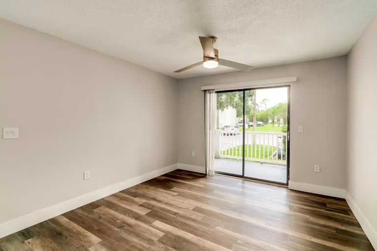 Empty room with hardwood flooring, beige walls, ceiling fan, and sliding glass doors opening to a deck with green railings
