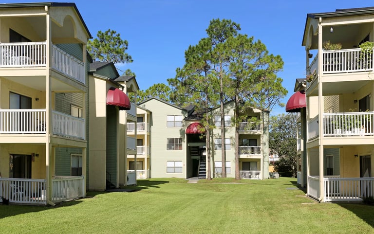 Residential apartment complex with multiple beige buildings featuring white railings and red awnings surrounding a green lawn