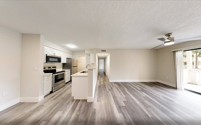 Modern open-concept apartment interior with kitchen, island counter, stainless steel appliances, wood-look flooring, and bright natural lighting