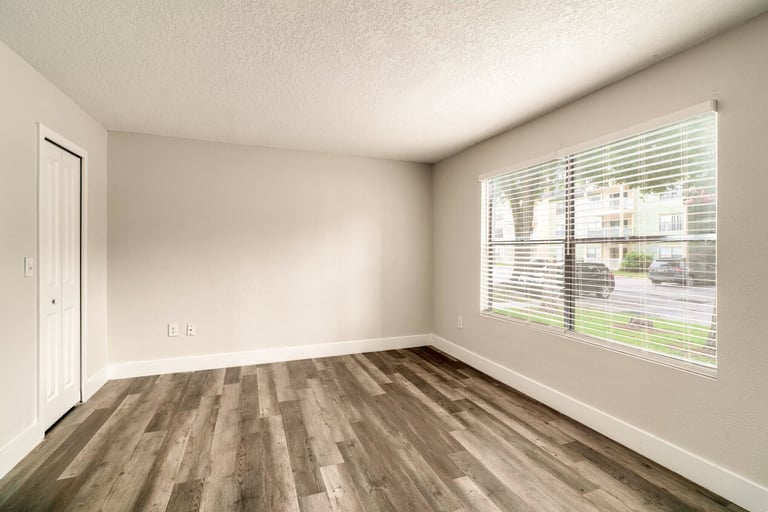Empty room with wood laminate flooring, white walls, door, and large window with blinds overlooking residential area