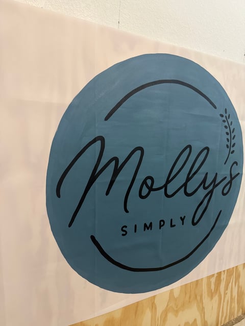 Blue circular sign with Mollys Simply logo and smiley face design on a white wall